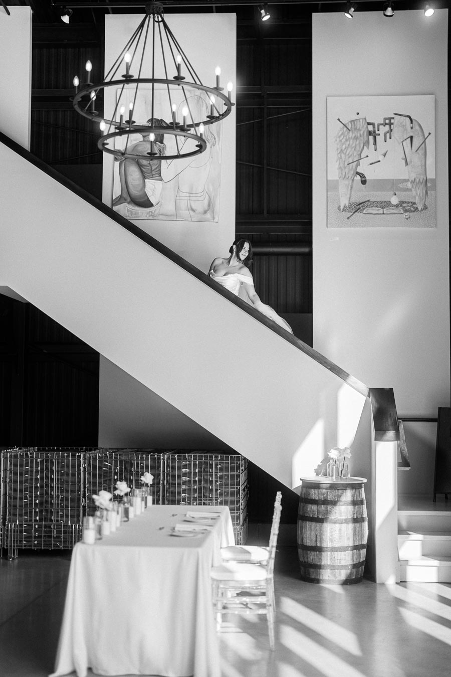 Black and white interior with a modern dining setup featuring a table draped in a white cloth, chairs, and floral arrangements beneath a staircase, art on walls, and a rustic chandelier.