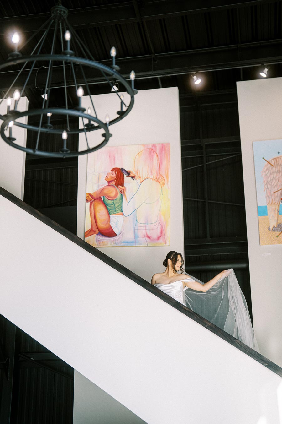 A woman in a white dress holding a sheer fabric stands on a staircase in an art gallery, with large colorful paintings in the background and a chandelier overhead.