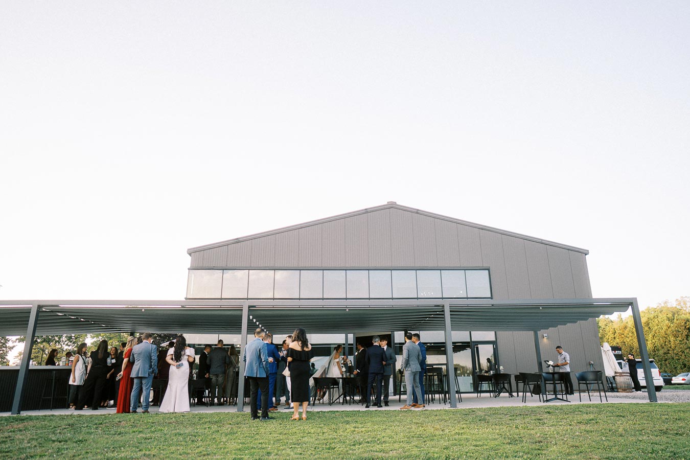 A group of people dressed in formal attire gather outdoors under a modern industrial-style building with a patio, possibly attending a wedding or social event, with a clear sky and grass lawn in the foreground.