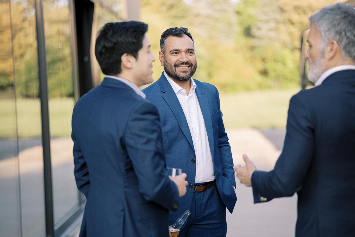 A group of three men in business suits engaging in a conversation outdoors, with one man holding a drink. The background shows a reflective glass wall and greenery, suggesting a professional networking event or social gathering.