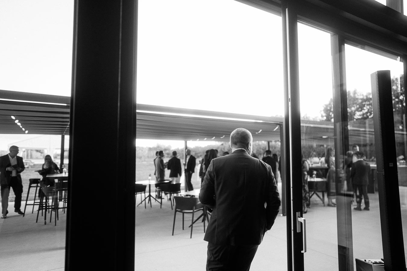 Black and white photo of a group of people in formal attire mingling at an outdoor event under a modern canopy, viewed through a glass doorway.