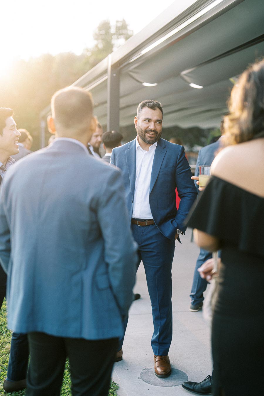 A group of people in formal attire socializing outdoors at an event, with a man in a blue suit smiling and engaged in conversation during sunset.