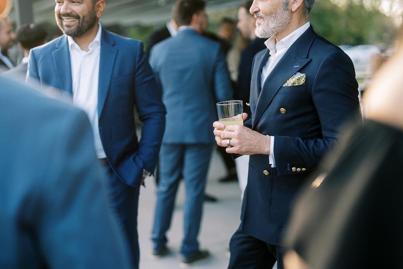 A group of professionals in stylish suits networking at an outdoor business event. A man in focus holds a drink and wears a navy suit with gold buttons, highlighting a sophisticated fashion sense.