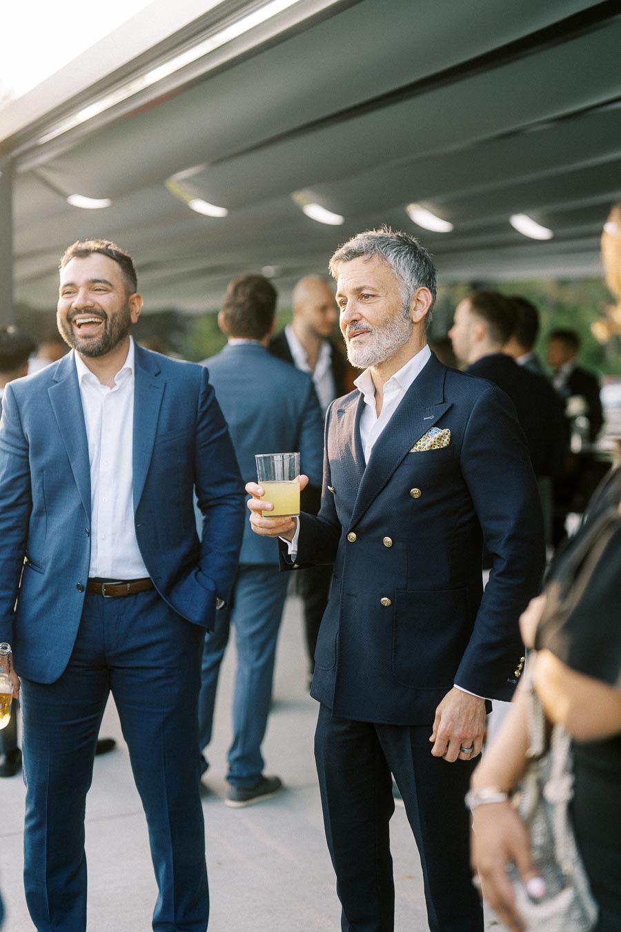 Two men in stylish suits enjoying a social gathering outdoors, one holding a drink and smiling, with a lively crowd in the background, creating a sophisticated and cheerful atmosphere.