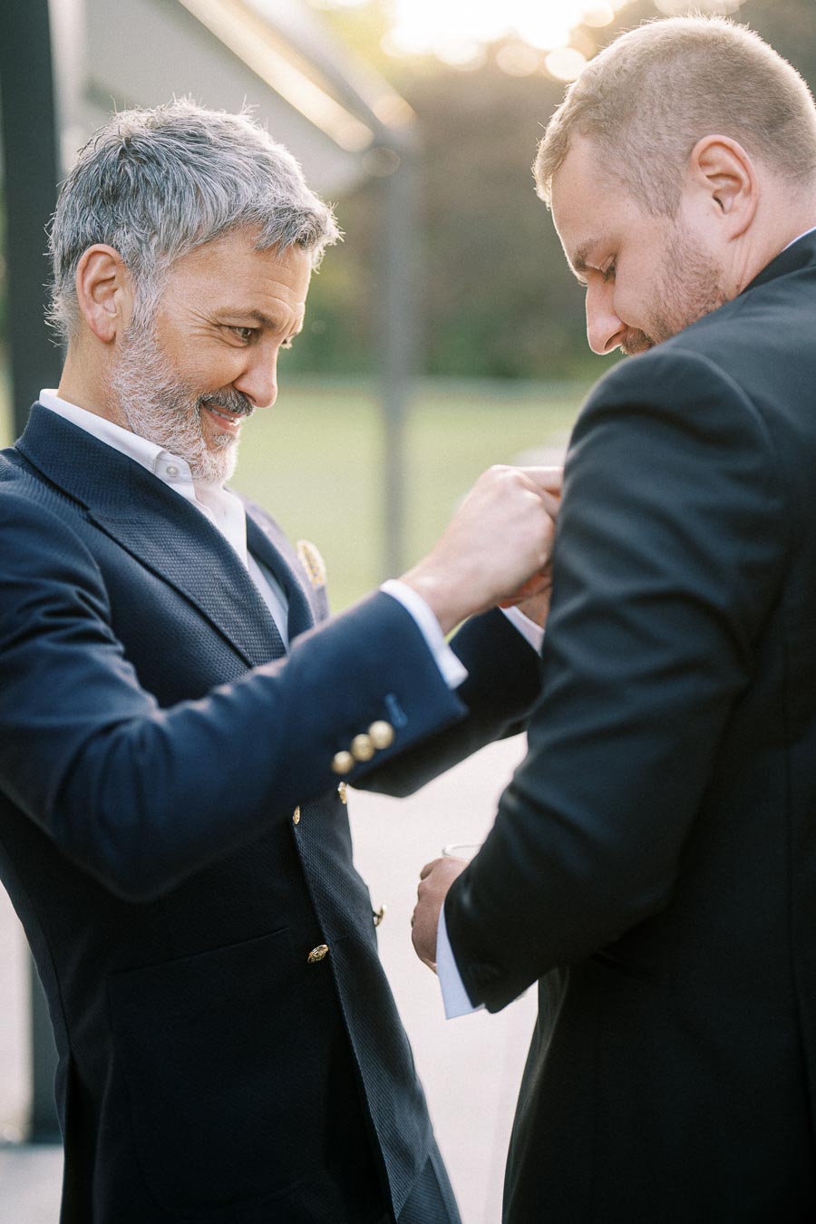 Two men in suits smiling and adjusting a boutonniere outdoors, showcasing elegant formal attire and capturing a joyful moment.