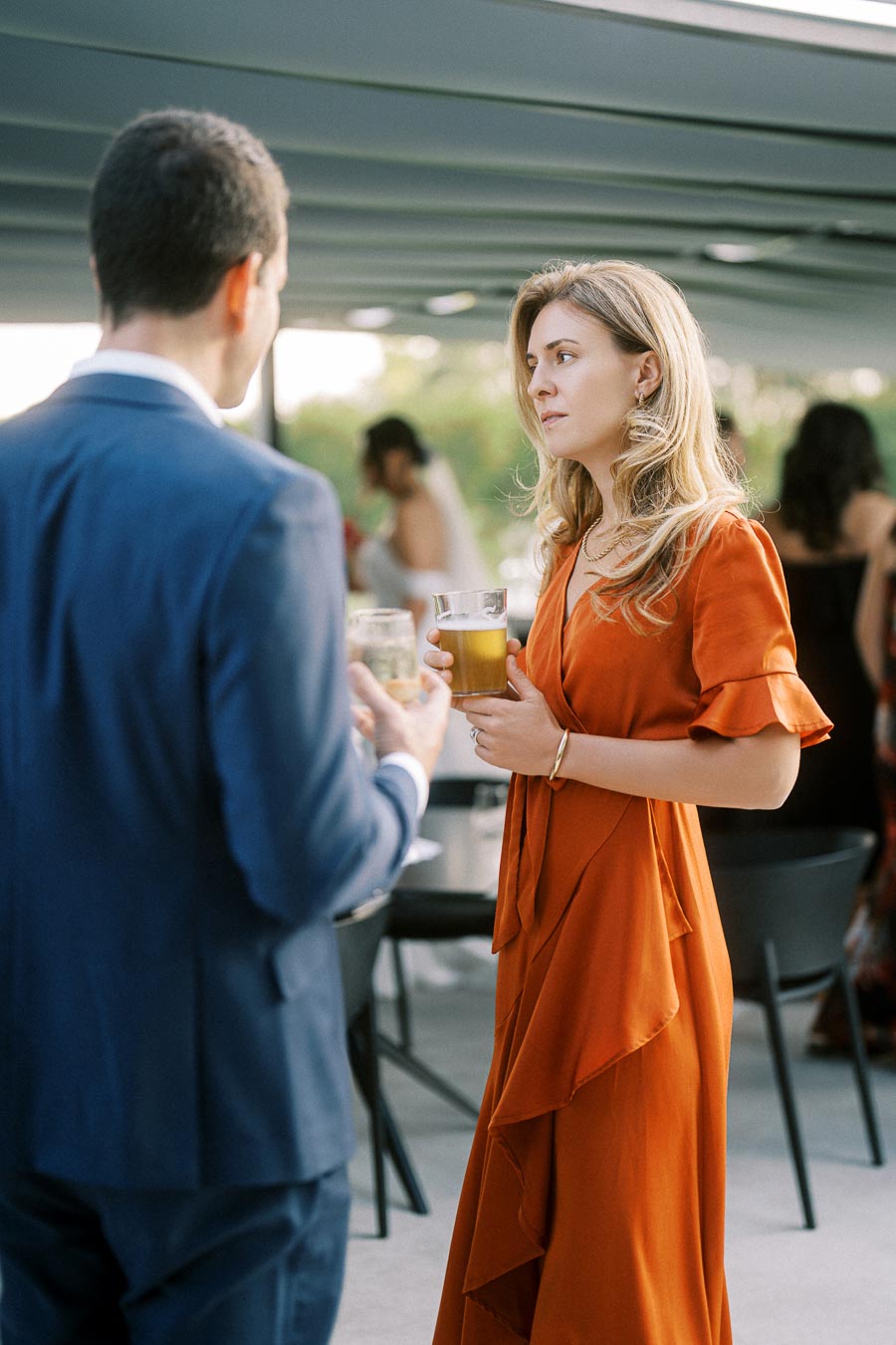 Elegant woman in an orange dress holding a drink while conversing at a formal outdoor event.