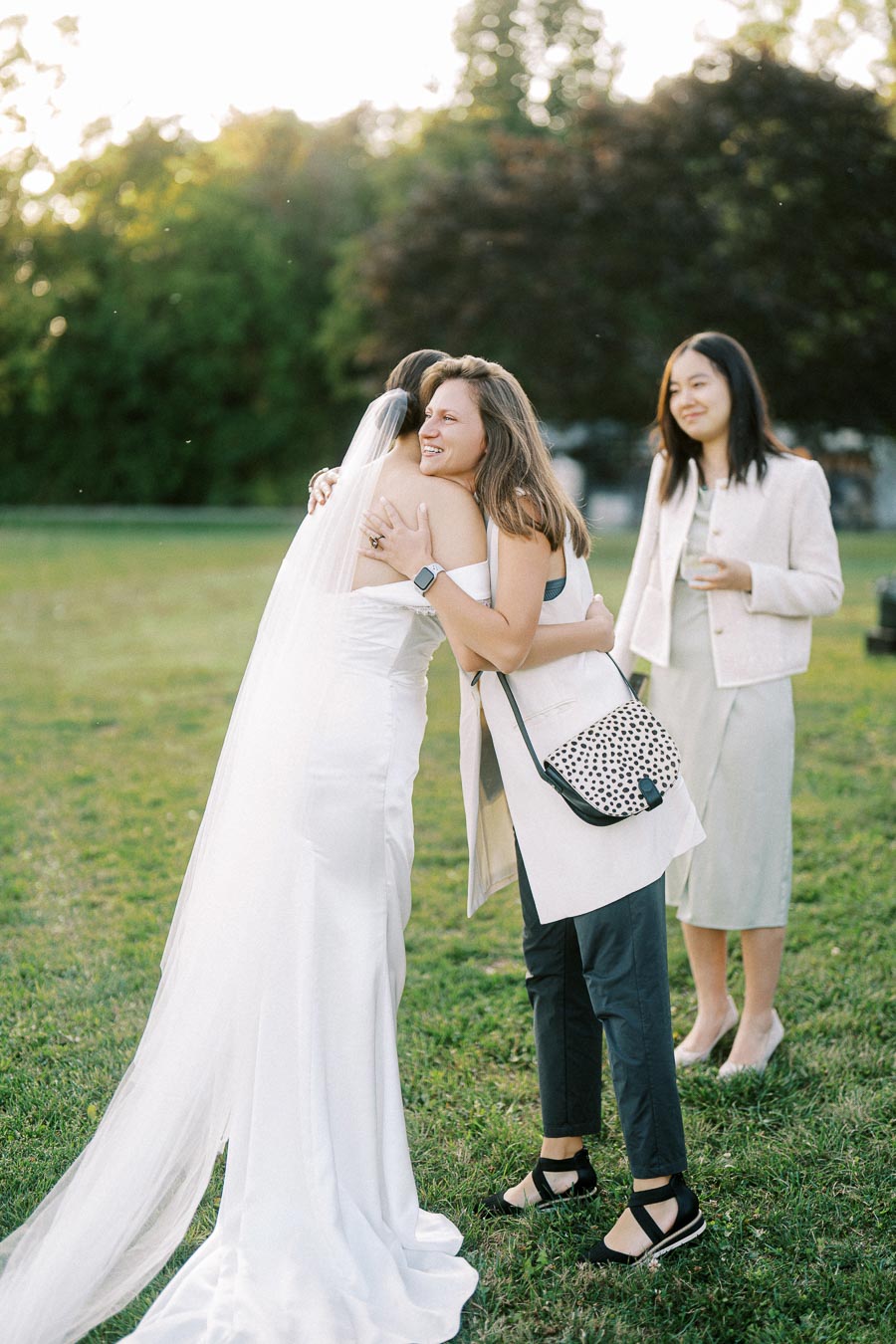 A bride in a white wedding gown with a long veil hugs a guest warmly at an outdoor wedding, while another guest smiles in the background. The scene is set in a grassy area with trees, capturing a moment of joy and celebration.
