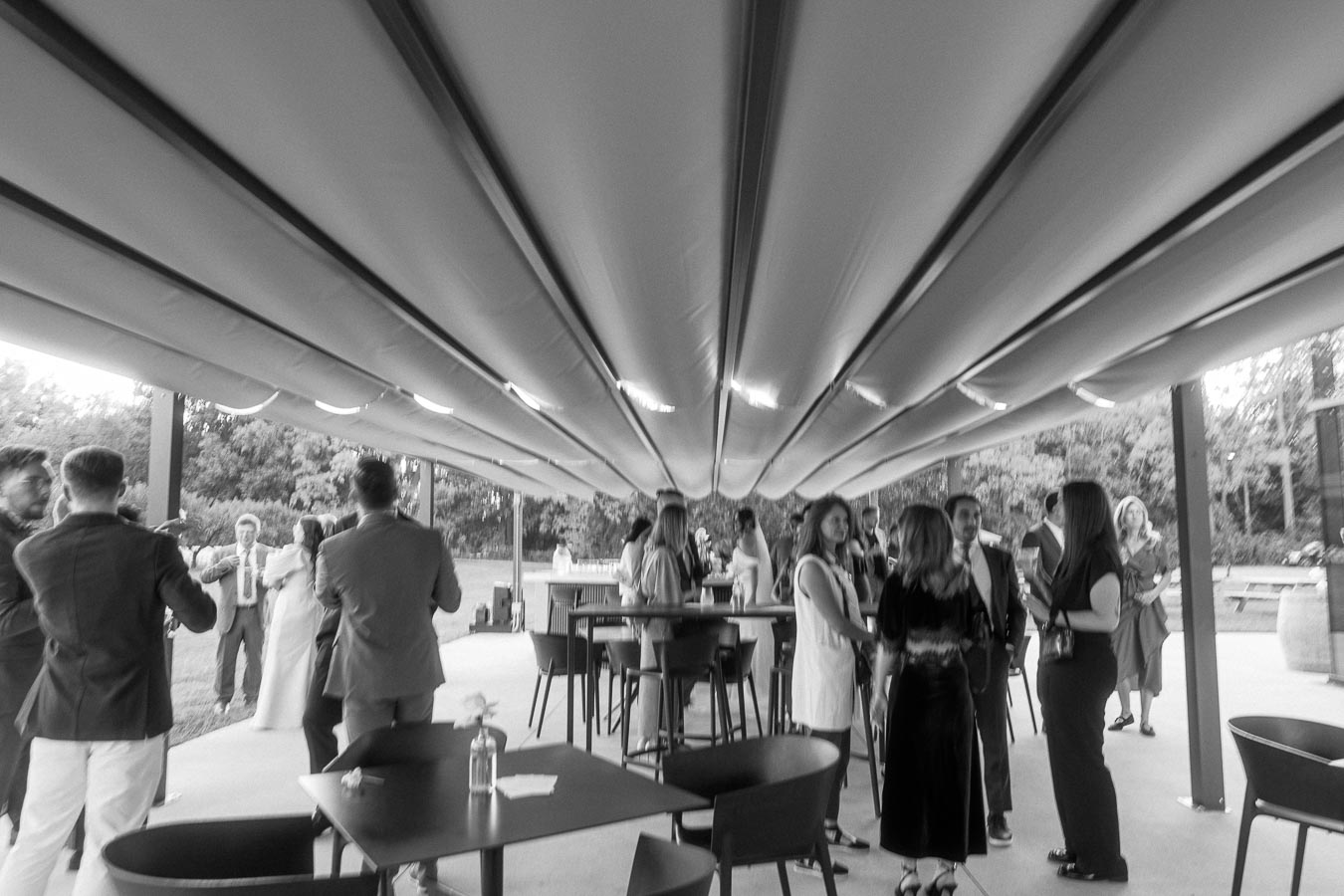 Black and white image of people mingling at an outdoor social event under a canopy, featuring tables and chairs, with trees visible in the background.