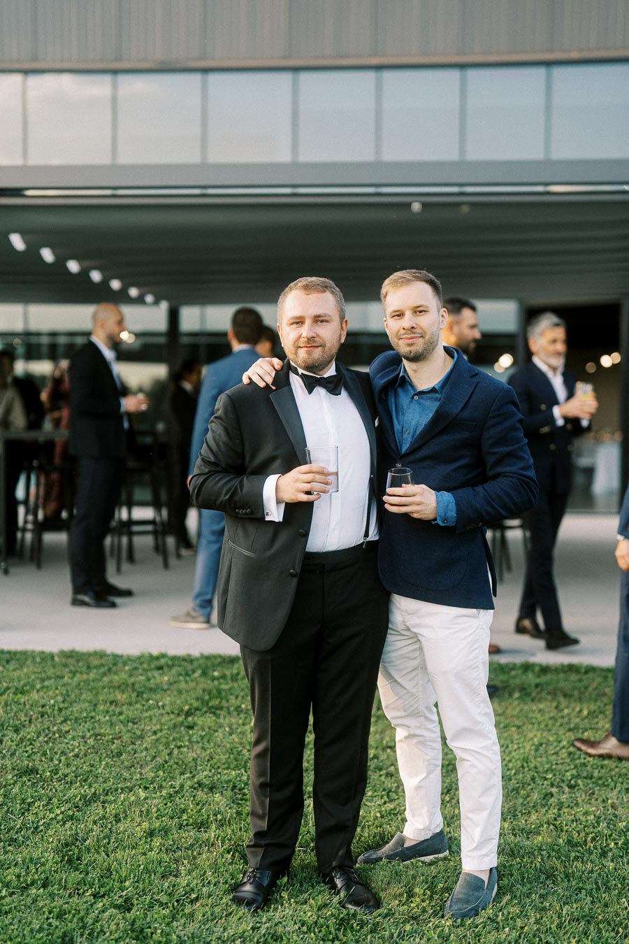 Two men dressed formally, one in a black tuxedo and the other in a blue jacket with white pants, smiling and holding drinks at an outdoor event with other attendees in the background.