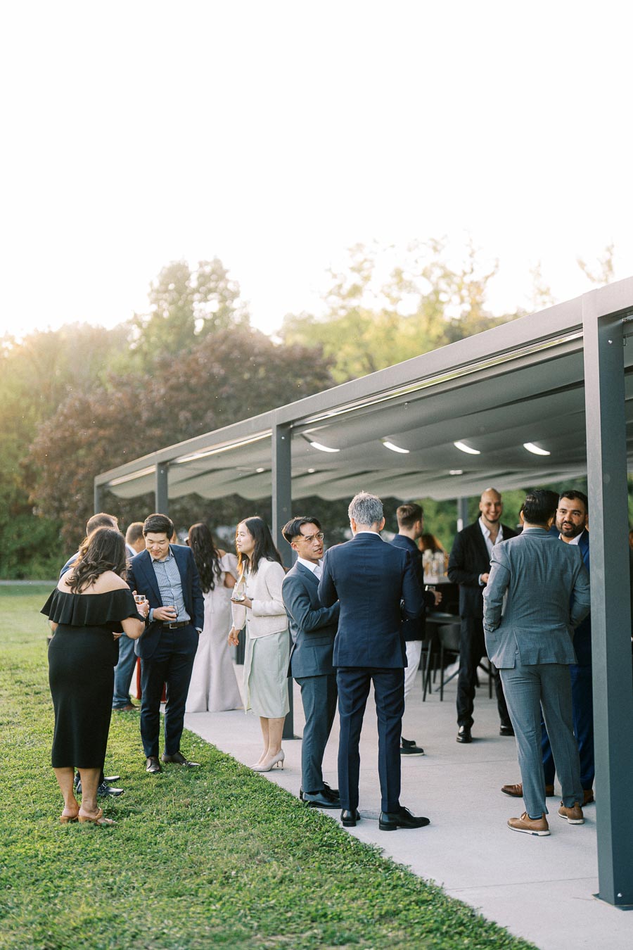 Outdoor social gathering with people in formal attire mingling near a modern canopy structure, set in a lush garden environment.