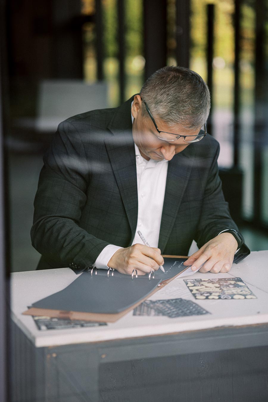 Man in a suit writing in a notebook at a desk in a modern office environment.