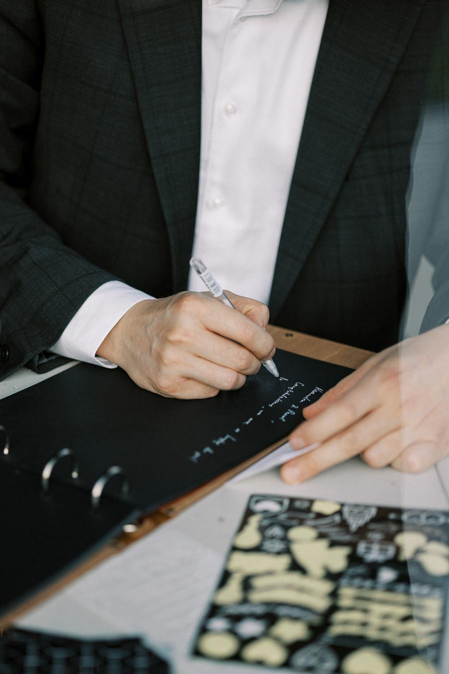 Person in a suit writing notes with a pen on a black notebook, featuring diagrams on a nearby page.