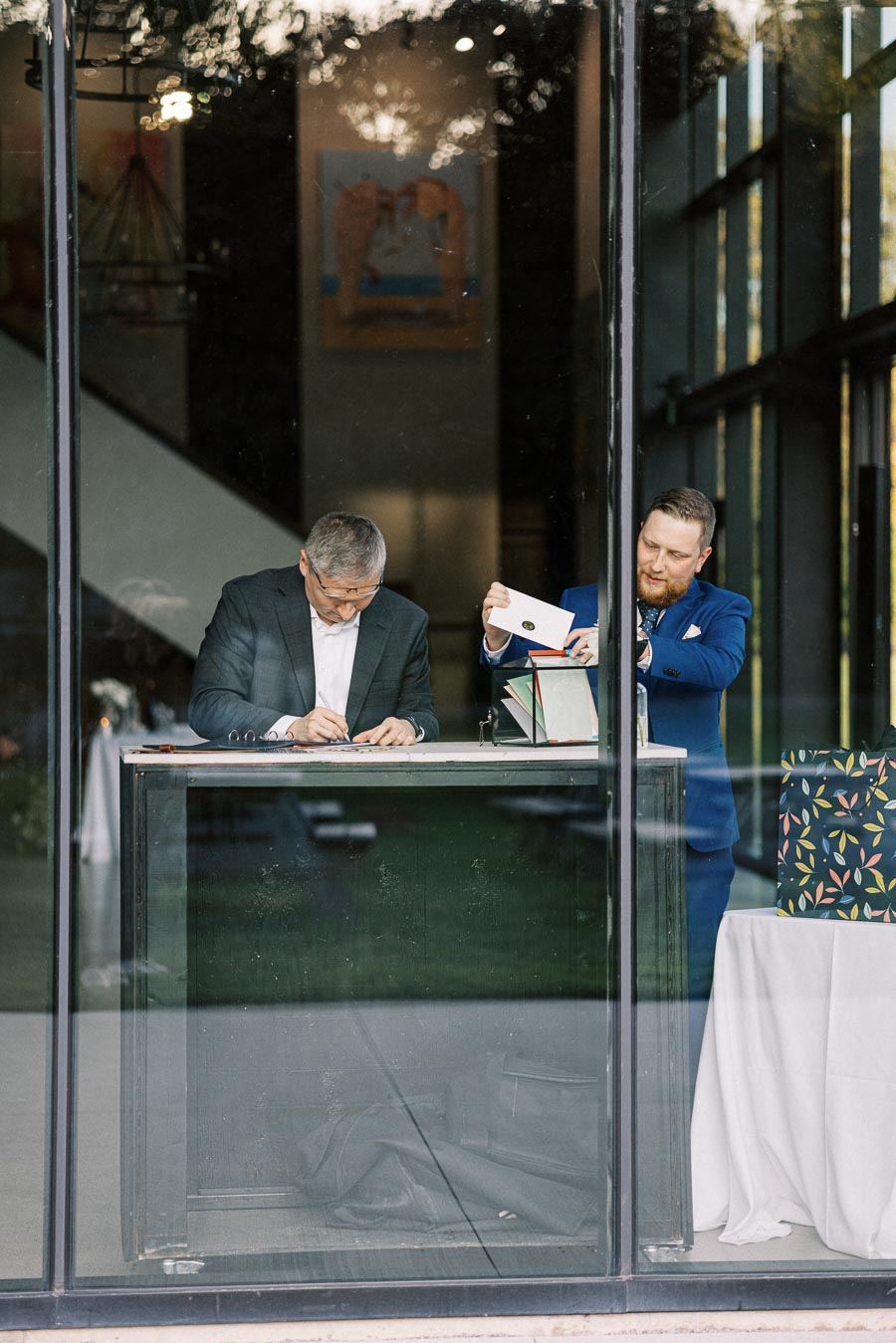 Two men in formal attire stand behind a glass window, with one signing a document and the other handling an envelope, in a modern office setting.