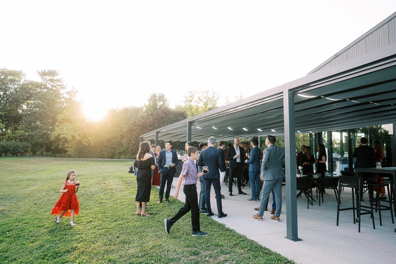 Outdoor gathering at sunset with people mingling under a modern pavilion, set beside a scenic lawn and lush greenery.