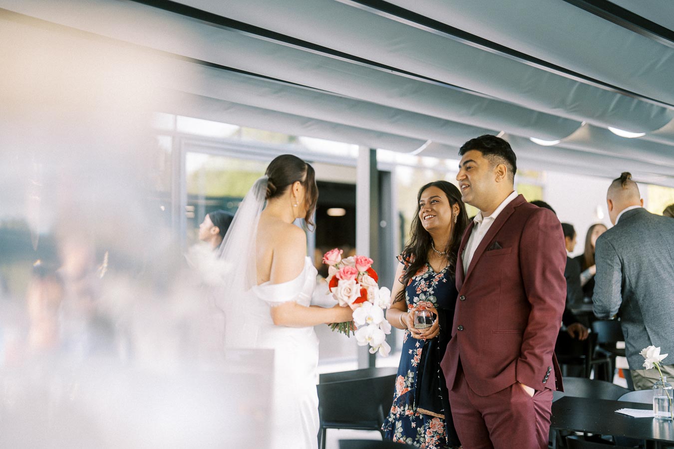 Wedding celebration scene with a bride holding a bouquet of red and white flowers, talking to guests in formal attire, under a modern, light-filled venue setting.