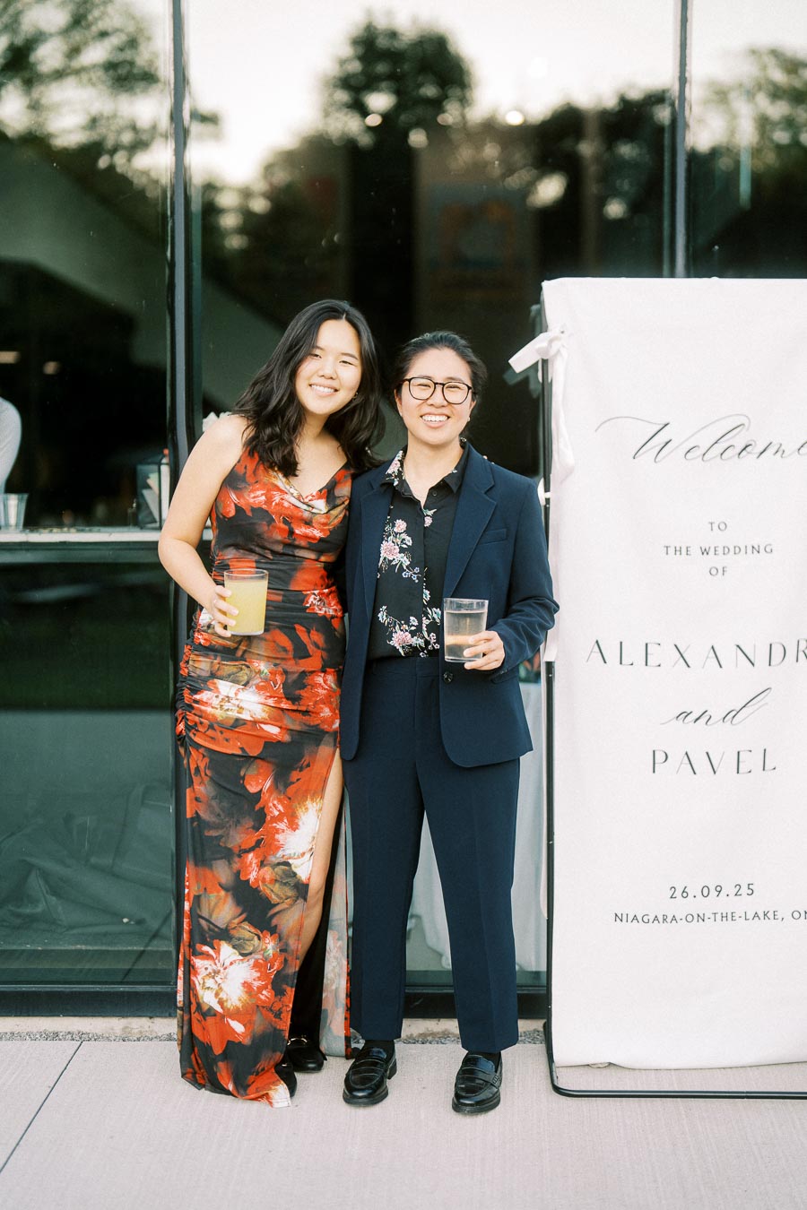 Two smiling guests posing at an outdoor wedding reception, one wearing a floral red dress and the other in a navy suit, standing next to a welcome sign for Alexandra and Pavel's wedding in Niagara-on-the-Lake.