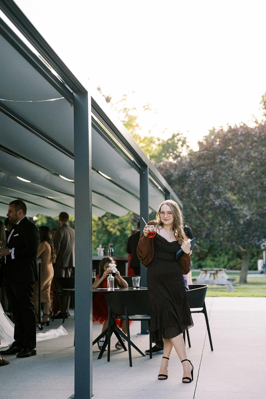 A woman in a black dress holding a drink and a clutch stands outside a modern venue with large glass panels, surrounded by people in formal attire. Trees and outdoor seating are visible in the background, suggesting a social event or wedding reception.