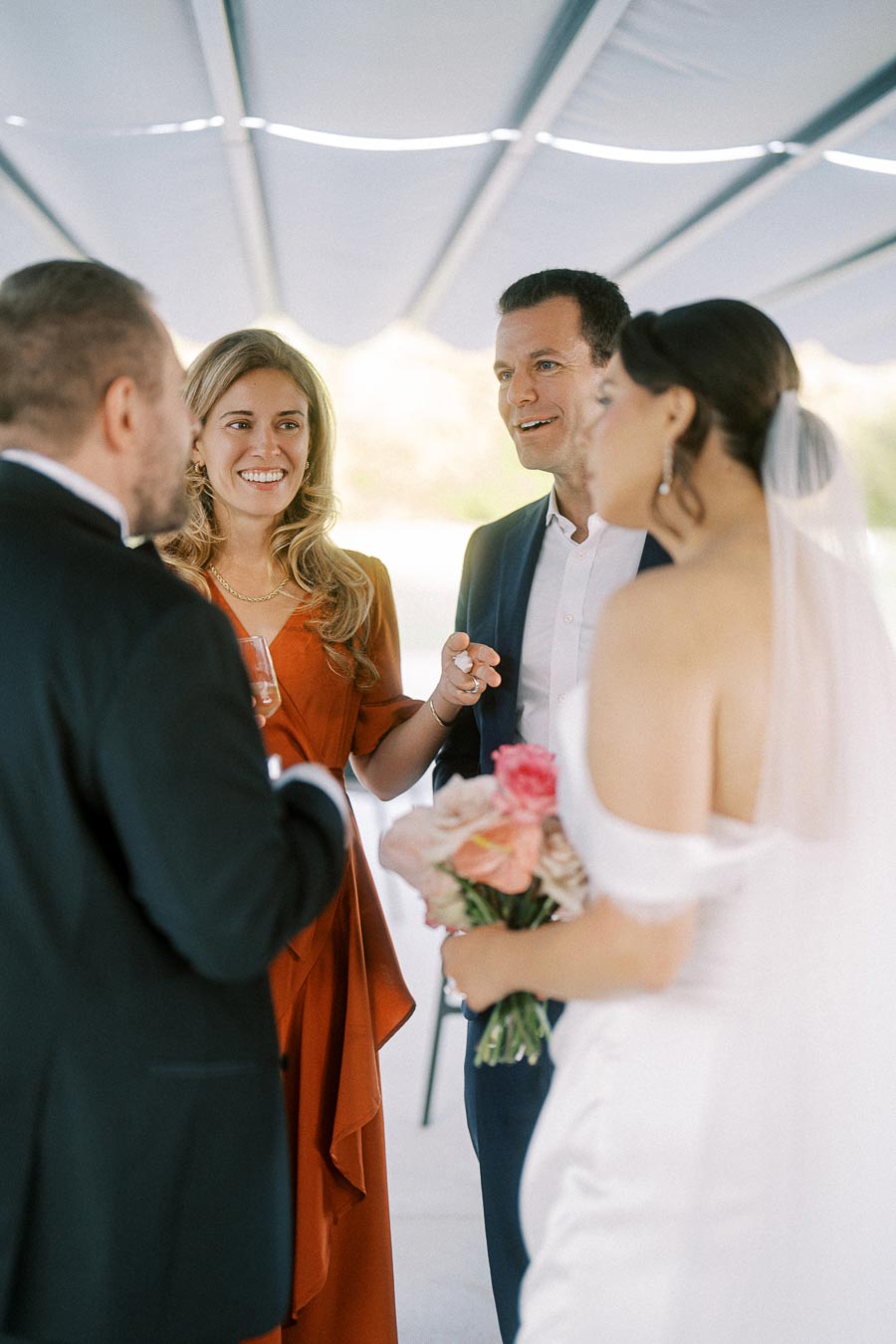 Wedding guests smiling and conversing under a tent as the bride, holding a bouquet, joins the group, dressed in a white gown, capturing a joyful moment at a celebration.