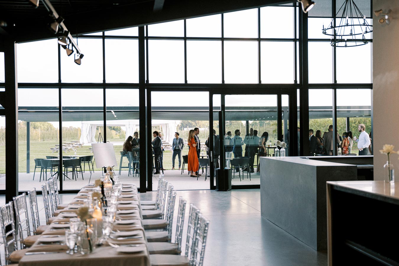 Luxurious event venue with elegant dining setup and guests mingling outside against a picturesque outdoor backdrop seen through large glass windows.