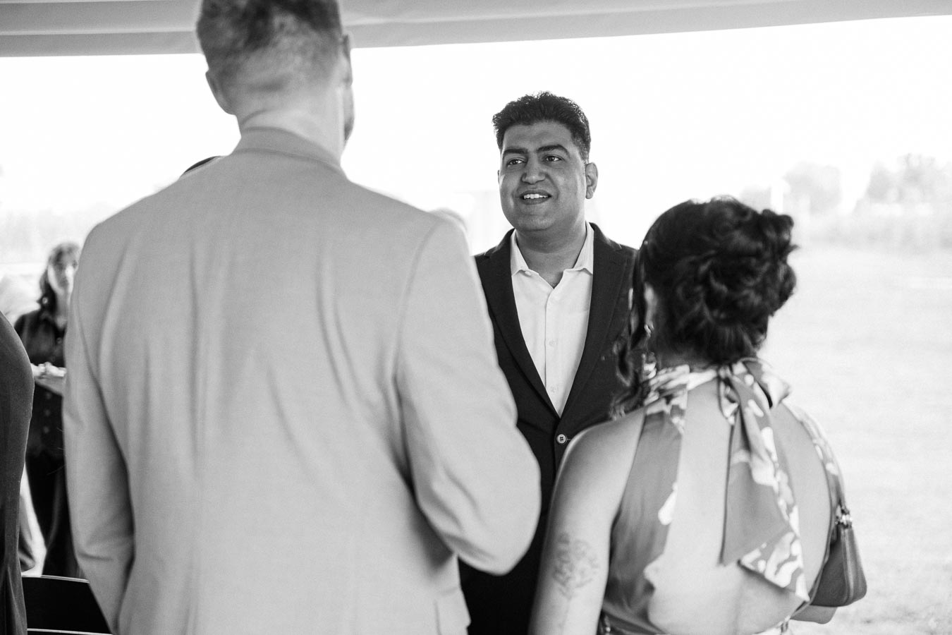 Black and white photo of three people in formal attire conversing at an outdoor event. The man facing the camera is smiling, while the other two have their backs to the camera. The setting appears casual yet elegant.