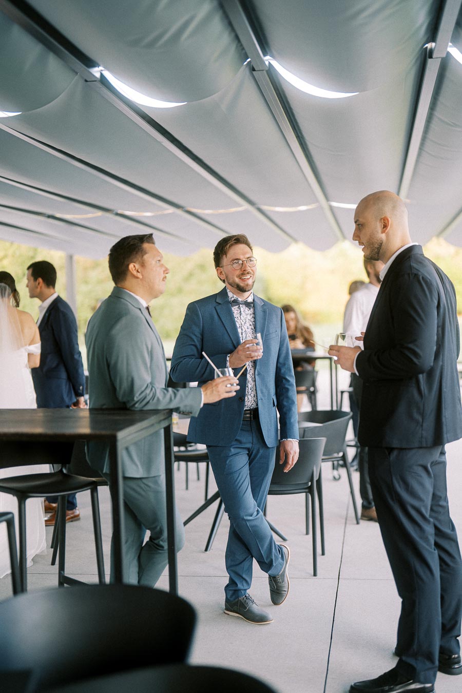 Three men in suits conversing and holding drinks at an outdoor event, with a bride and groom visible in the background.