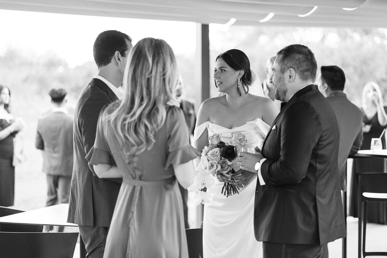 Black and white photo of a wedding reception, featuring a bride holding a bouquet of flowers, engaged in conversation with three guests, all elegantly dressed.