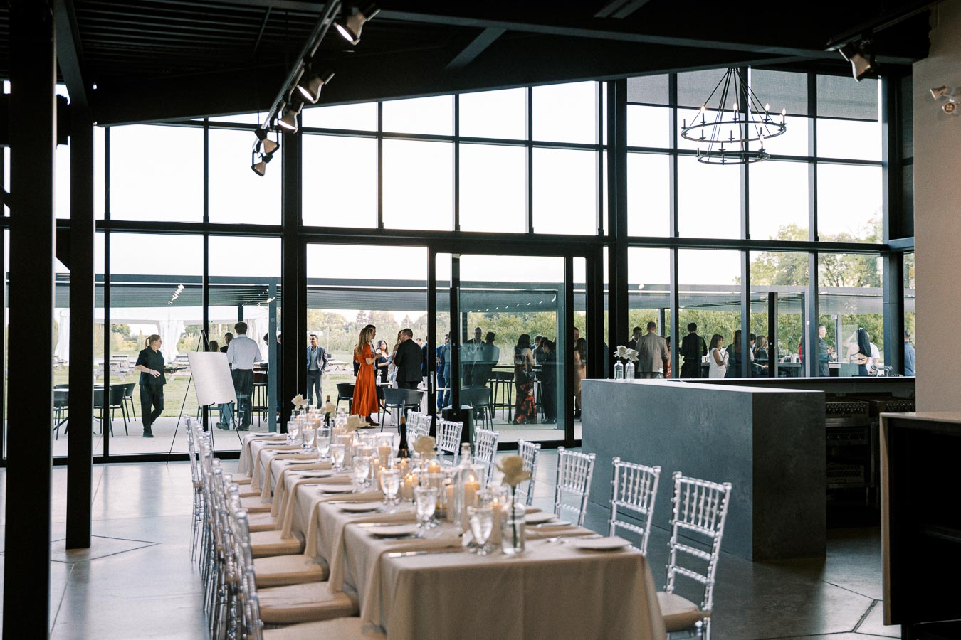 Elegant modern banquet hall with a long table set for a formal event, featuring white linens, candles, and glassware. Large windows offer a view of an outdoor gathering, showcasing attendees mingling at a stylish event venue.