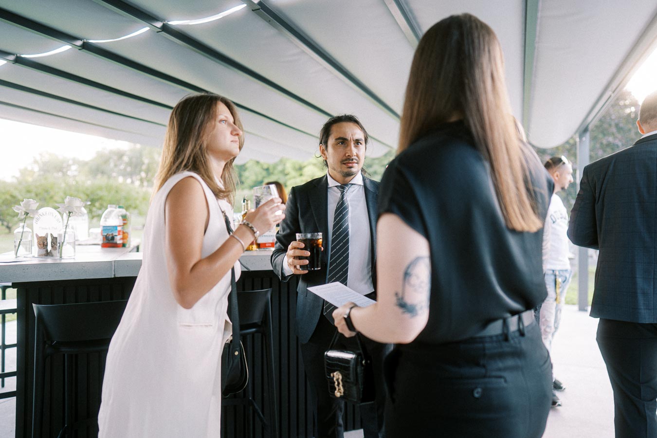 Group of people socializing at an outdoor event under a canopy, with drinks in hand and informal attire.
