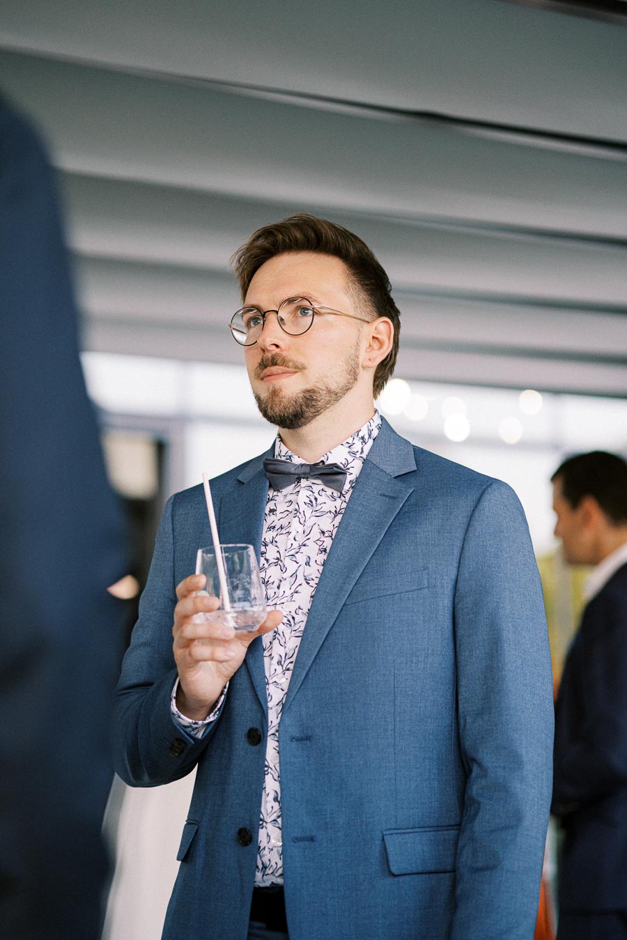 A man in a blue suit and floral shirt holding a glass with a straw at a formal event.