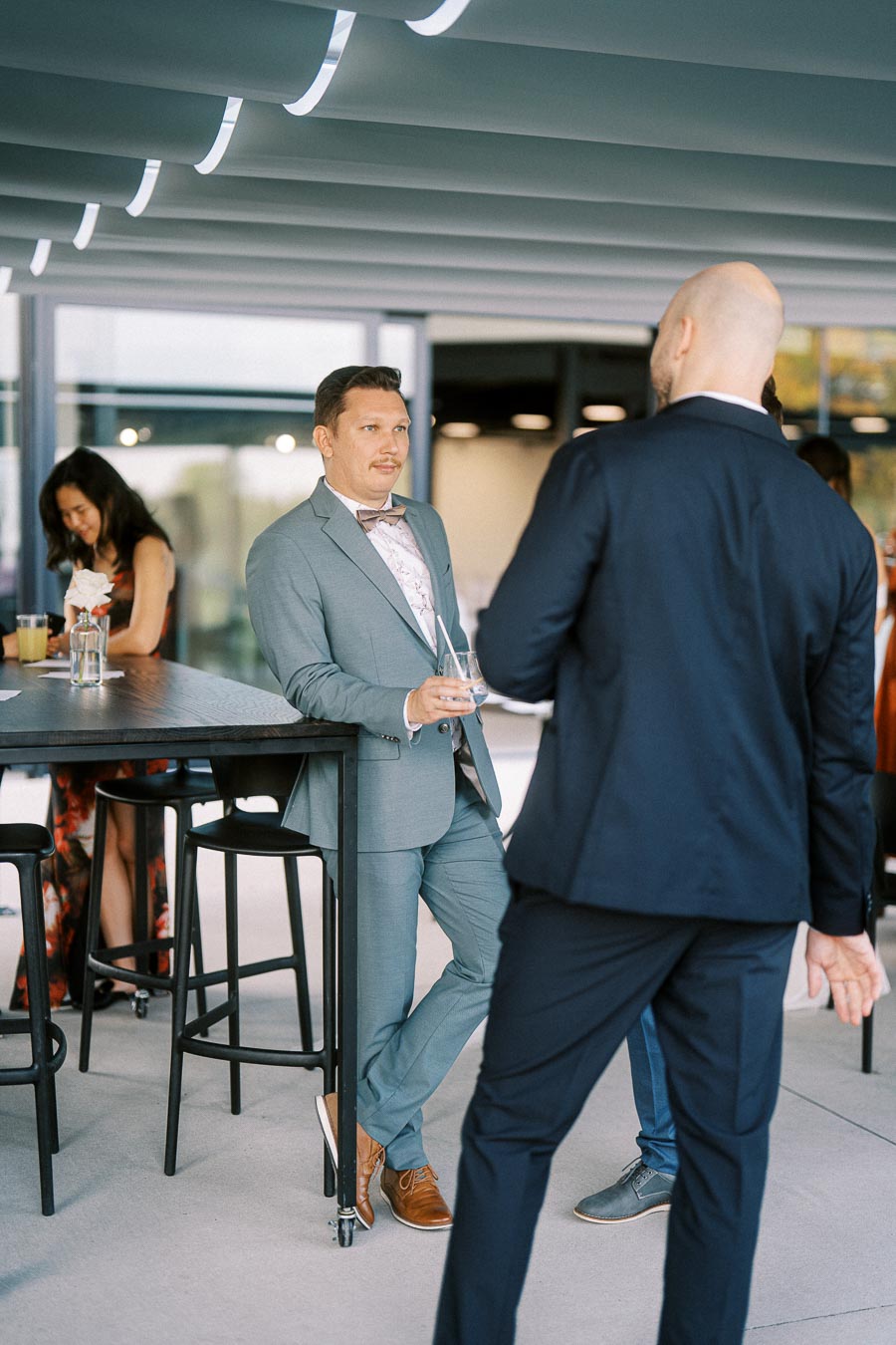 Group of elegantly dressed individuals socializing at an outdoor event, with one person in a gray suit holding a drink and leaning against a table, creating a relaxed and sophisticated atmosphere.