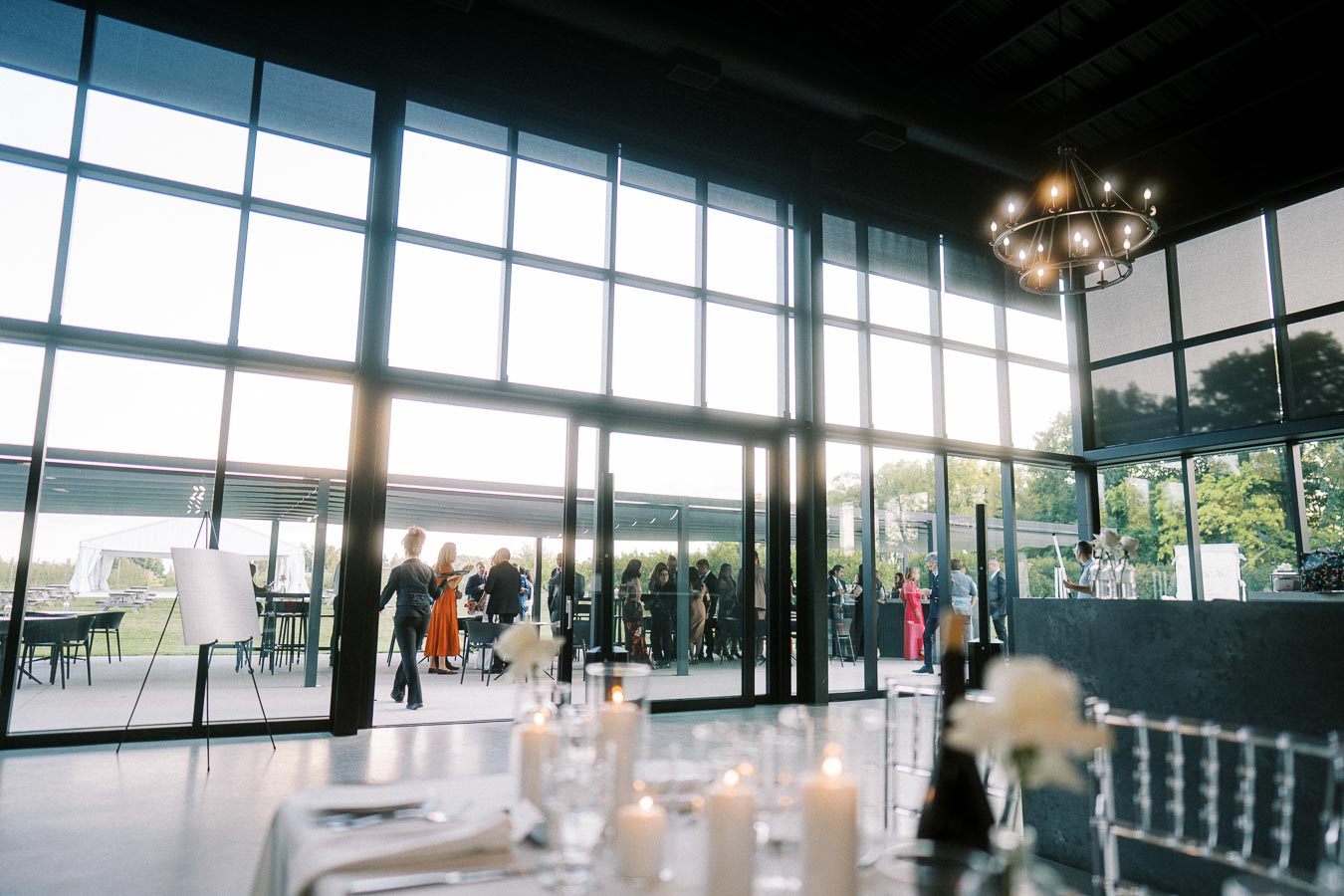 Elegant indoor event with large floor-to-ceiling windows showcasing a gathering of people outside, featuring a modern chandelier and candlelit dining setup inside.