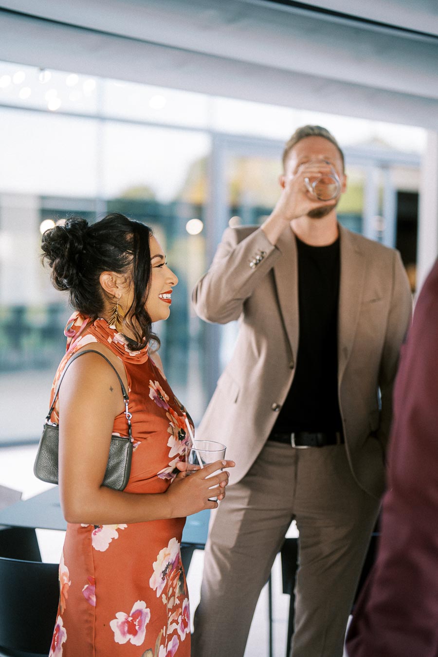 A woman in a floral dress with a small handbag smiles as she converses with a man in a beige suit drinking from a glass at a social gathering.