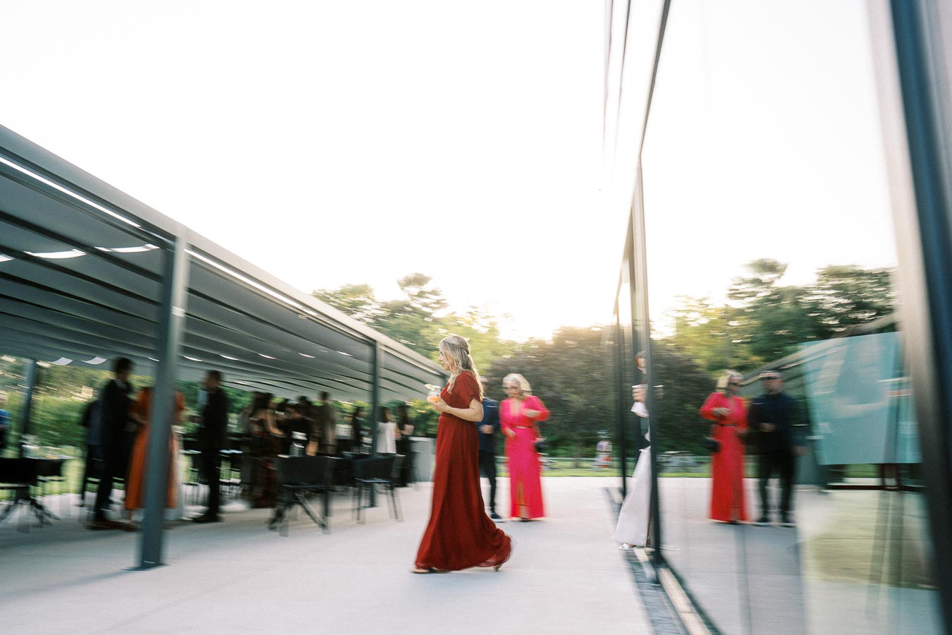 A woman in a red dress walks past a modern glass building during an outdoor event, with other elegantly dressed individuals gathered in the background under a canopy.