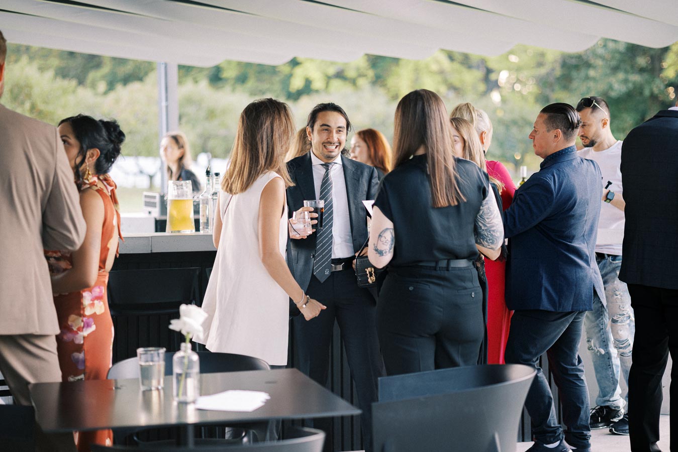 A group of people interacting at an outdoor social event near a bar, with individuals dressed in a mix of formal and casual attire, surrounded by greenery in the background.
