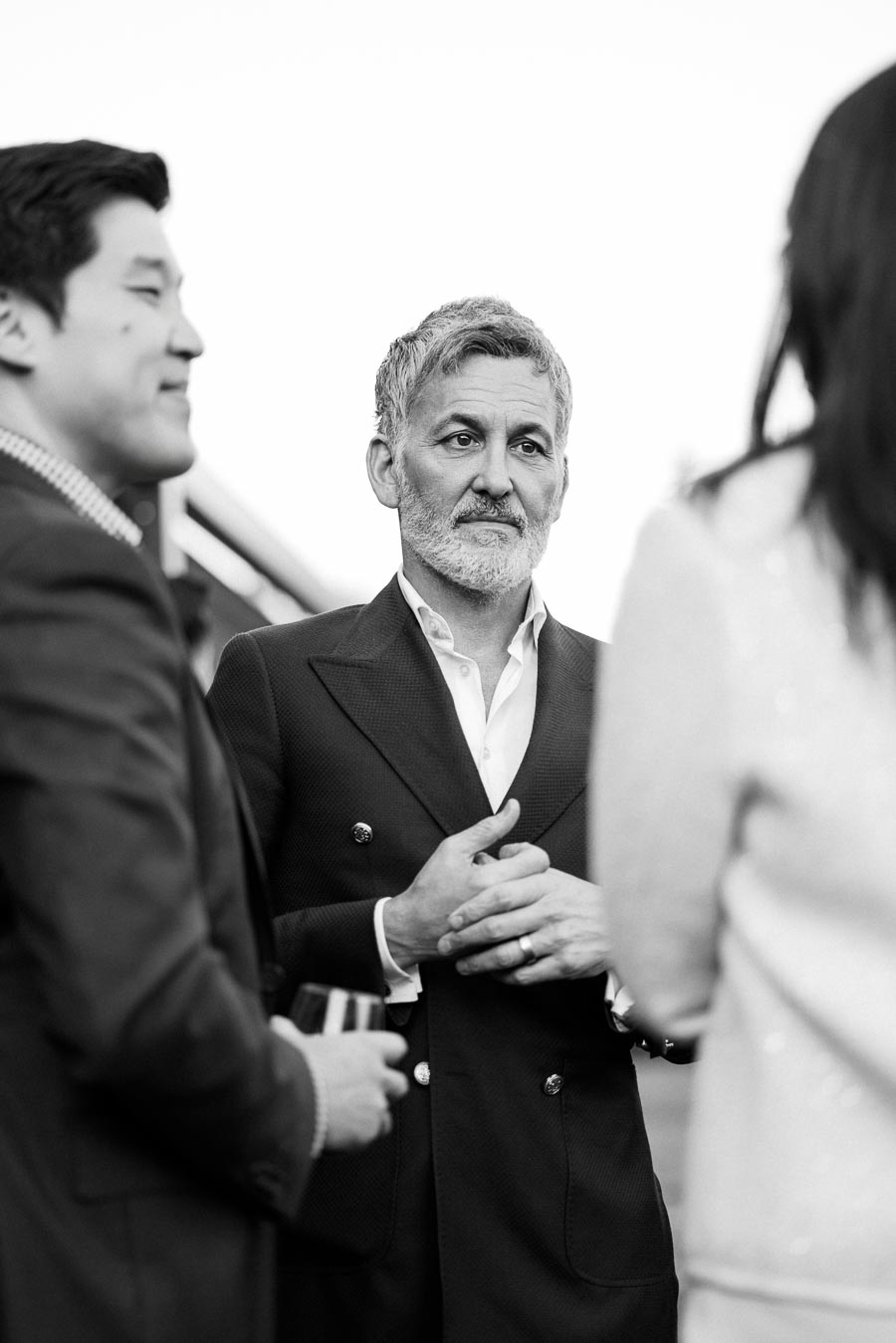 Black and white image of a distinguished older man with gray hair and beard, wearing a formal suit, standing among a small group of people at a social gathering.