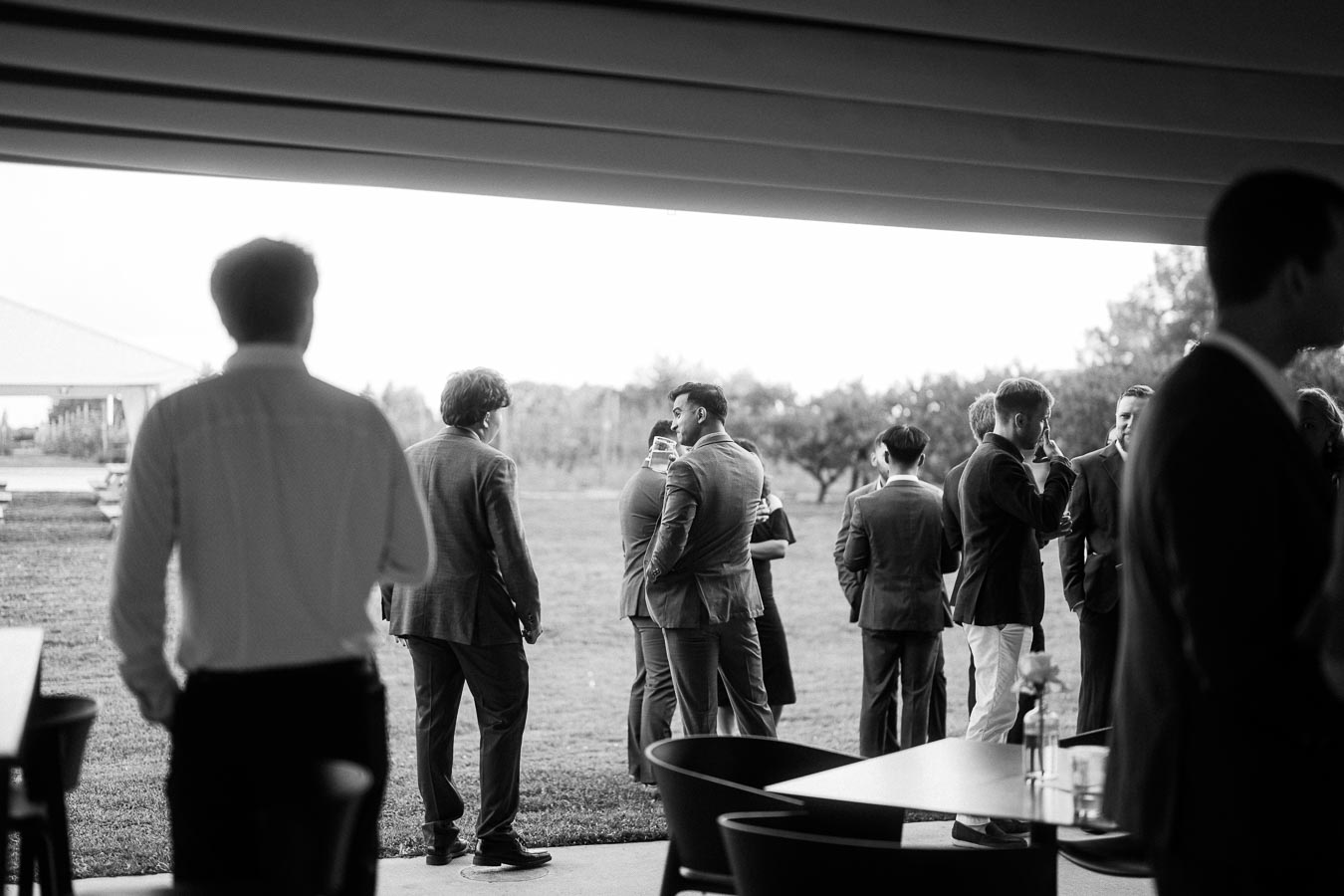 Black and white photo of a group of people in suits socializing at an outdoor event, with a grassy field and trees in the background.