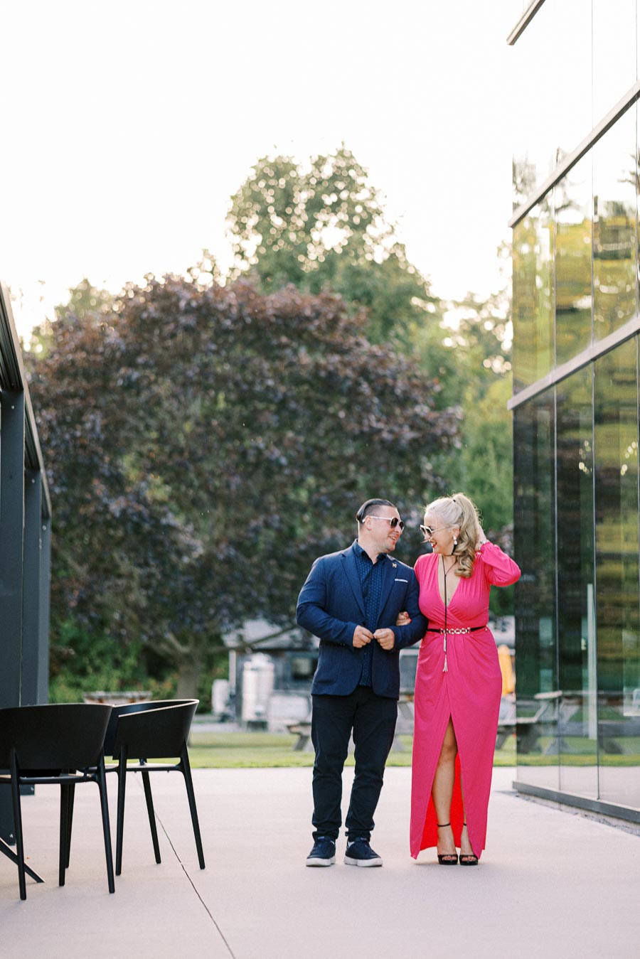 A stylish couple walks arm in arm outside, with the man in a blue suit and sunglasses and the woman in a vibrant pink dress. They are smiling at each other against a backdrop of greenery and modern architecture.