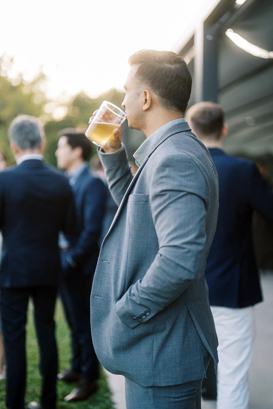 A man in a gray suit enjoys a drink outdoors at a social gathering, with other people in formal attire in the background. The scene is well-lit, suggesting early evening or late afternoon.