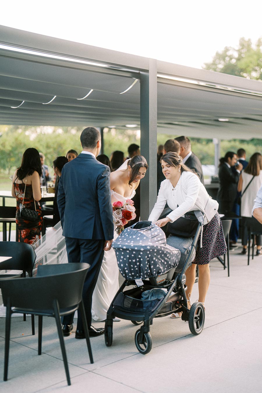 Wedding reception gathering with a bride holding a bouquet, guests chatting, and a woman attending to a baby stroller.