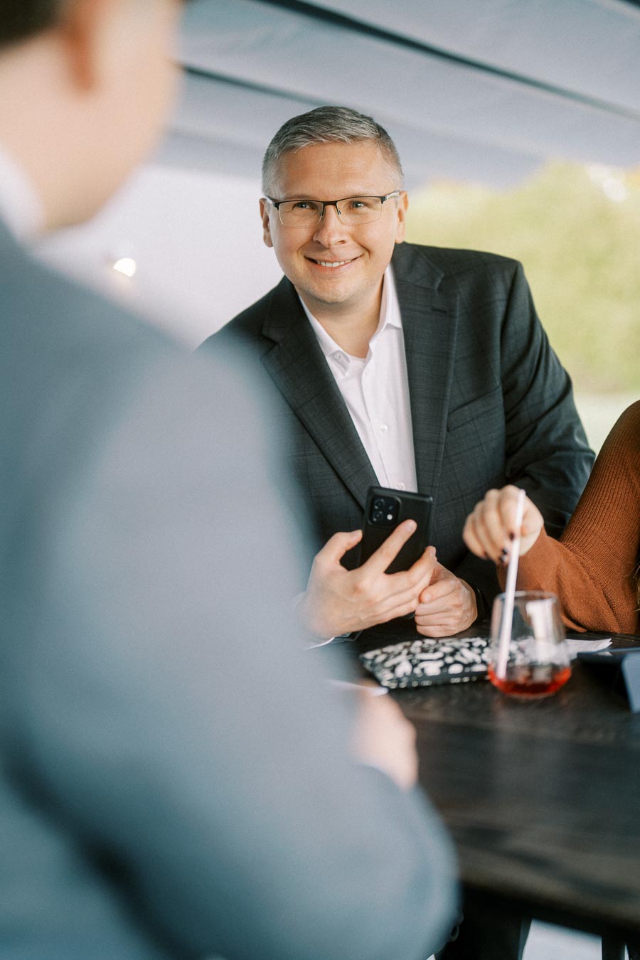 Man in glasses and suit smiling while holding a smartphone during a casual business meeting, with a blurred figure in the foreground and a glass of red drink nearby.