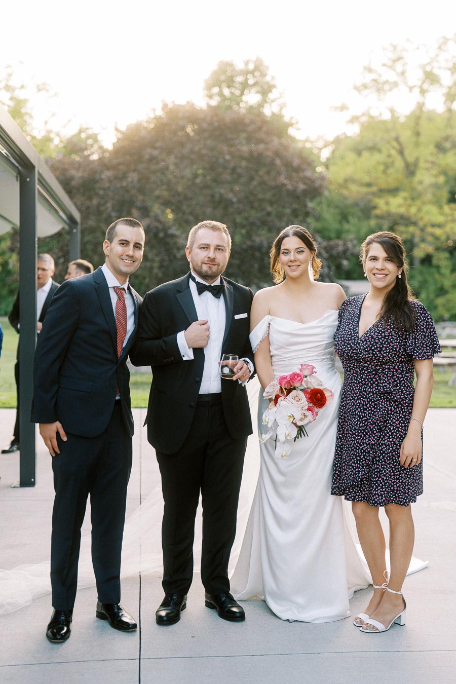 A group of four people at an outdoor wedding celebration, with two men in formal suits and two women in elegant dresses, the bride holding a bouquet of pink and white flowers, smiling against a backdrop of greenery.
