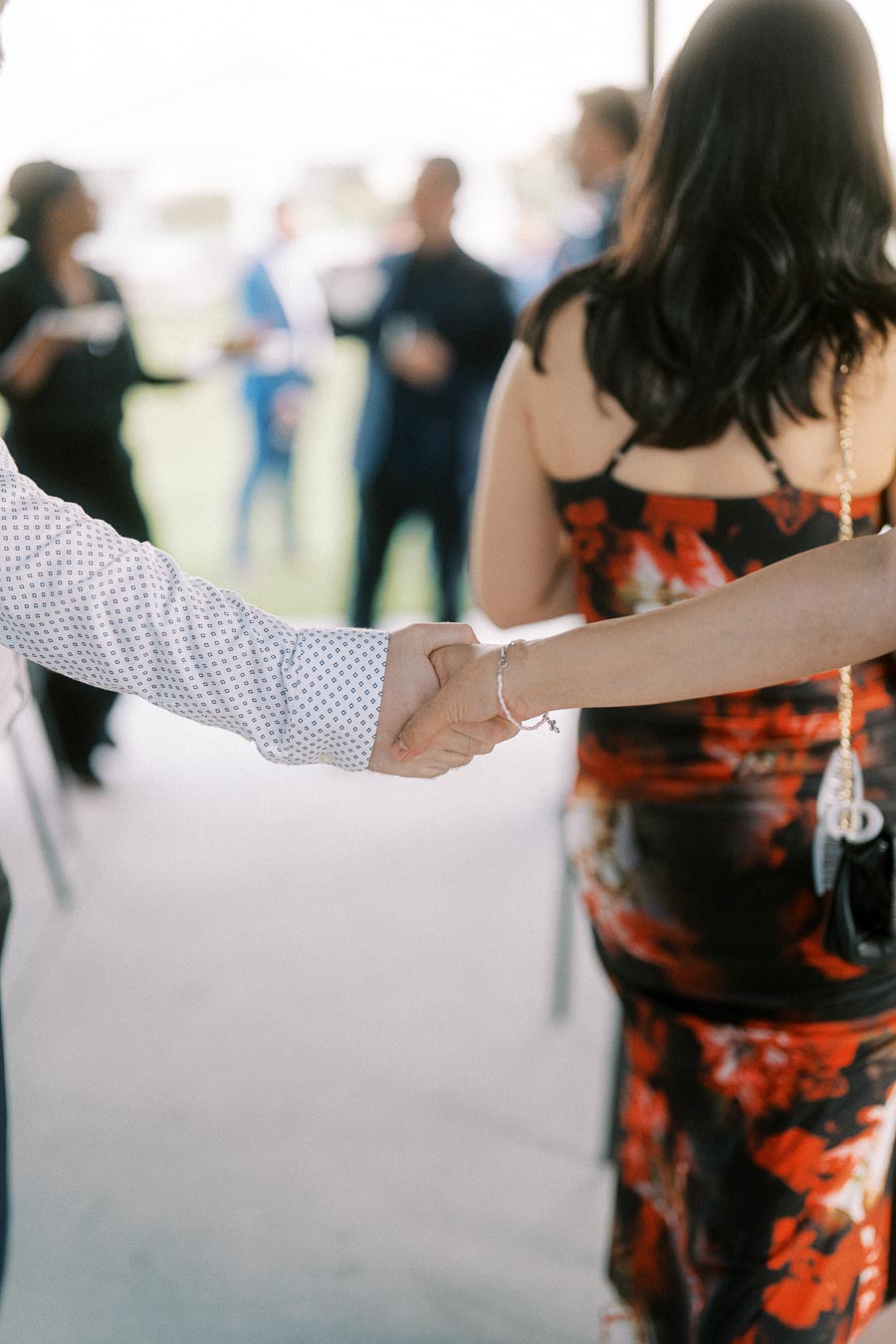 Two people shaking hands at an outdoor event, with a blurred background of people socializing, dressed in formal attire.