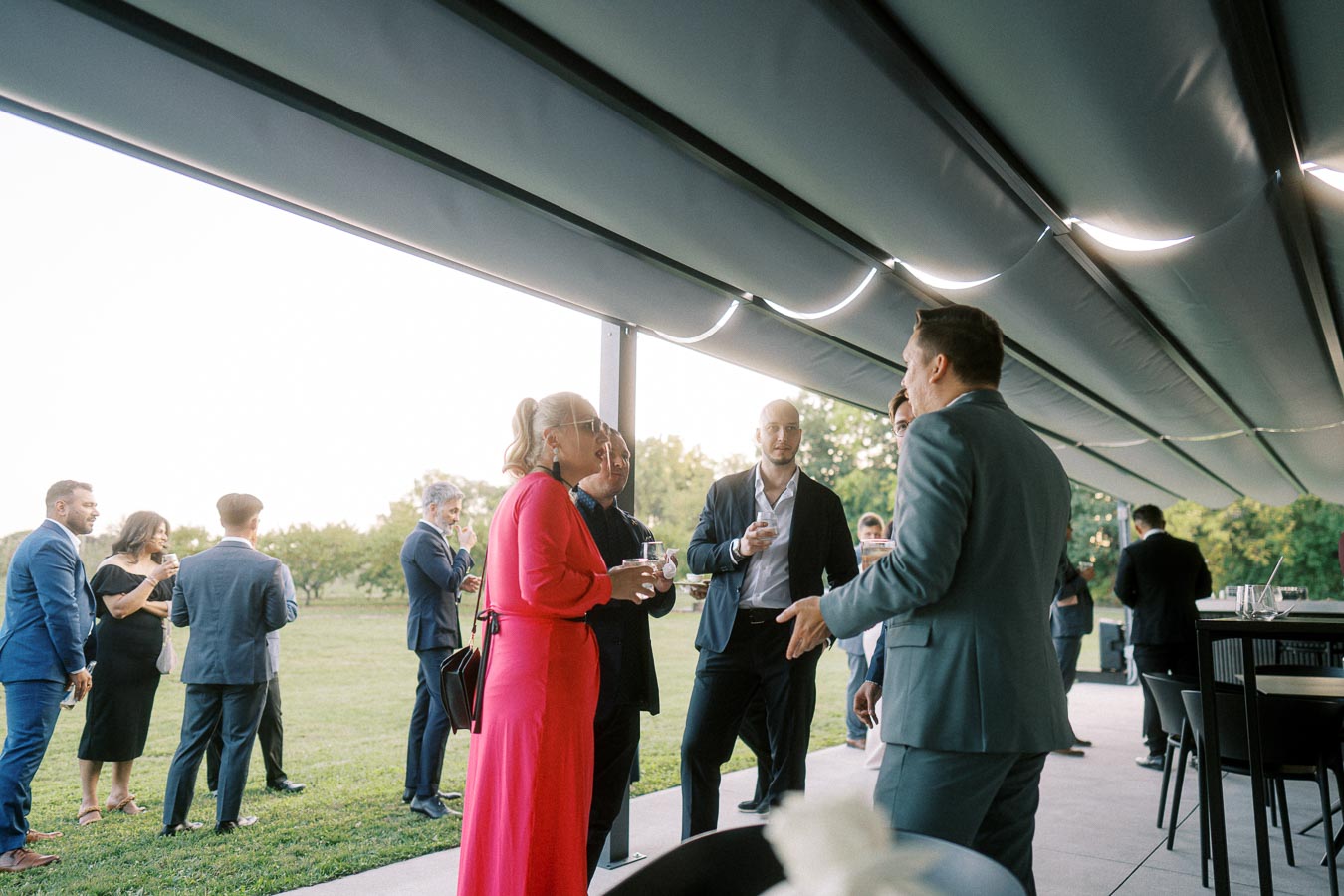 Outdoor business networking event with professionally dressed people engaging in conversation under a modern canopy, set against a backdrop of lush greenery.