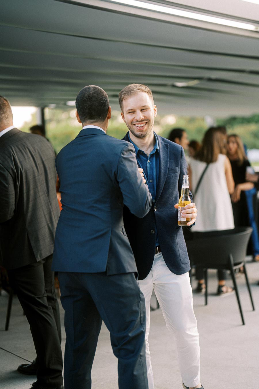Two men in suits shaking hands and smiling at an outdoor social event, one holding a drink, with a group of people in the background.