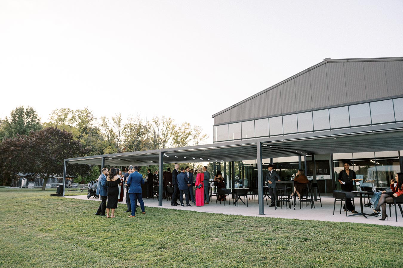 Outdoor social gathering at a modern event venue, featuring guests socializing under a covered patio, surrounded by trees and greenery.