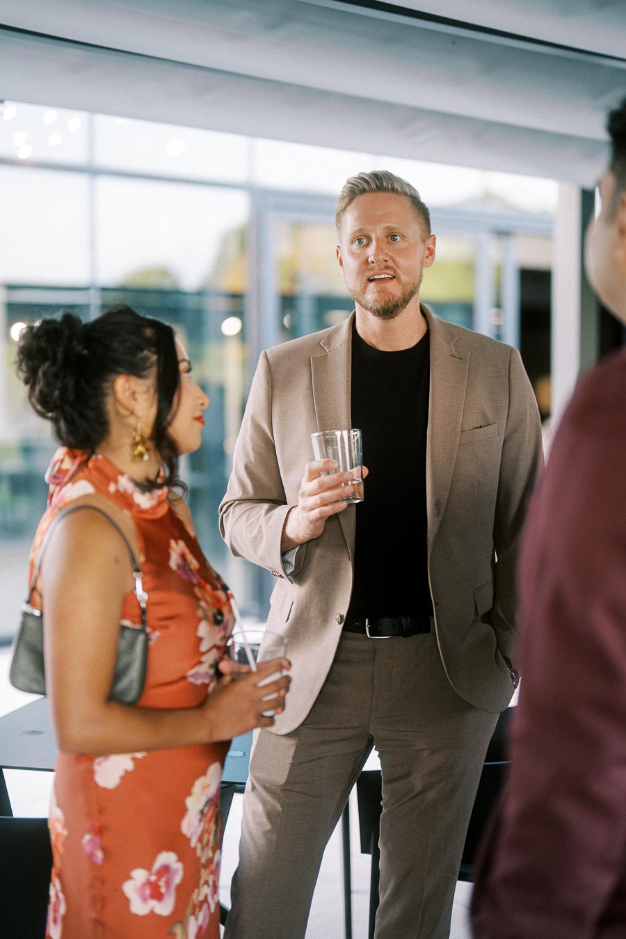 Group of people engaged in conversation at a social event, featuring a man in a beige suit holding a drink and a woman in a floral dress, with modern indoor setting.