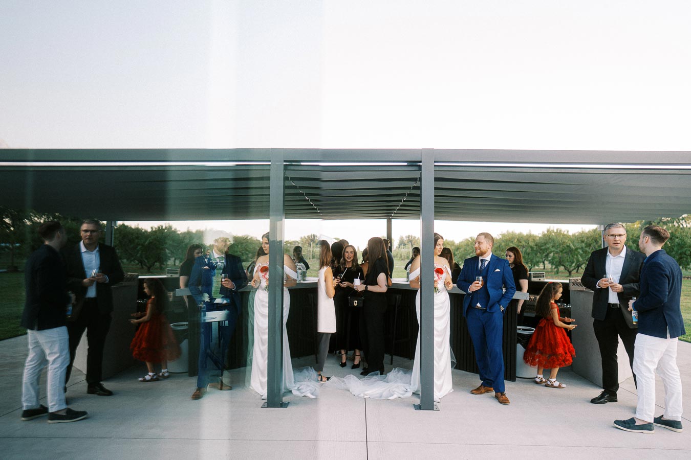 Outdoor wedding reception with guests socializing under a modern pergola. A bride in a white dress holds a bouquet, while guests in formal attire enjoy drinks and conversations.