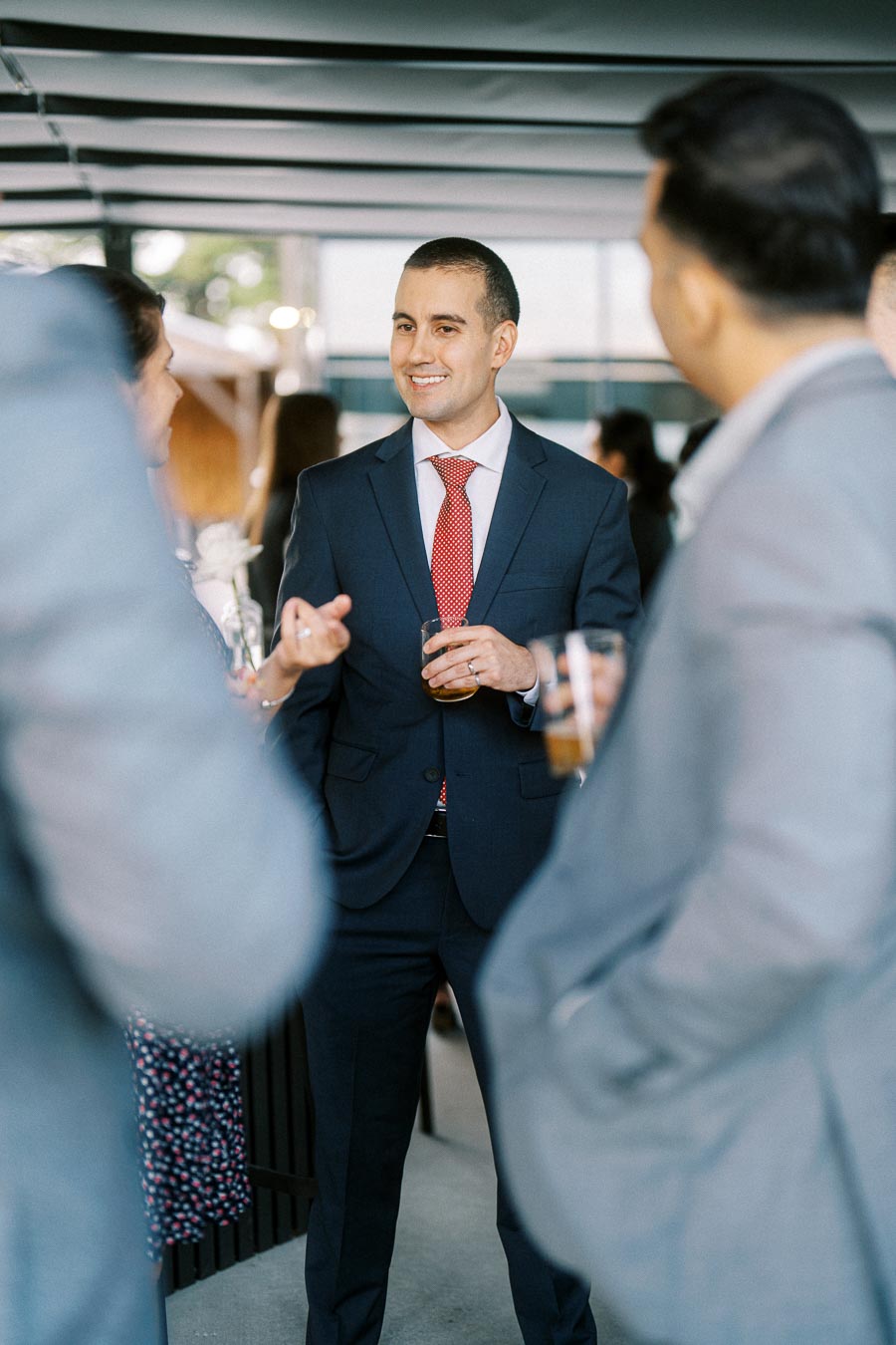 A man in a suit and red tie converses with colleagues at a professional networking event, holding a drink and smiling, with a blurred background of other attendees.
