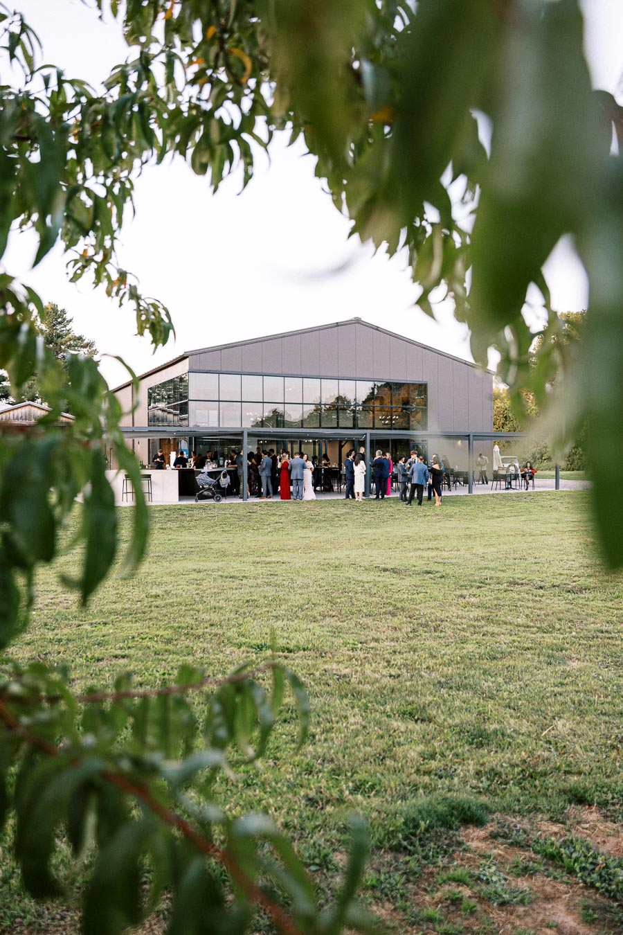Modern barn-style event venue with glass windows hosting an outdoor gathering, surrounded by lush greenery and framed by tree leaves.