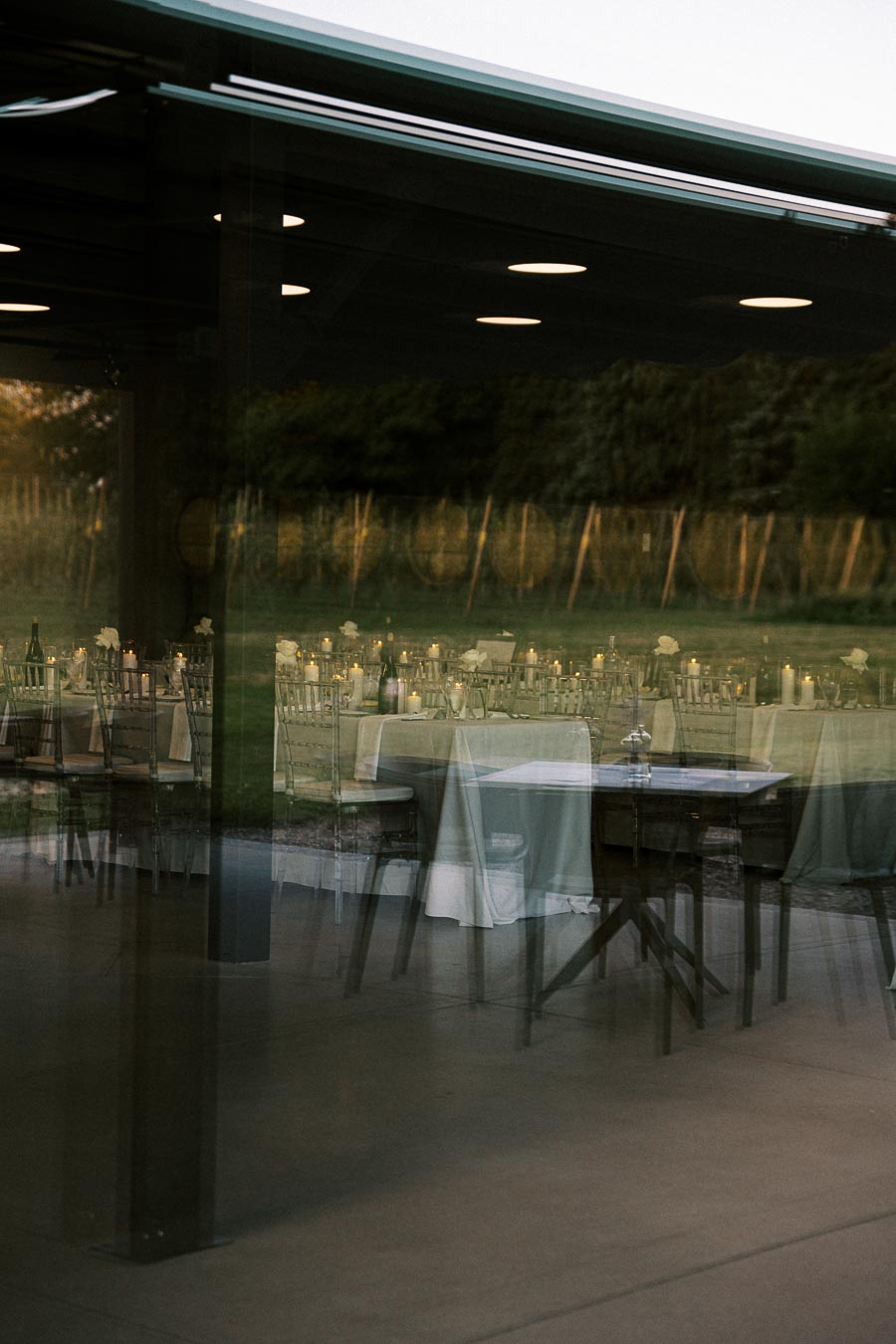 Elegant outdoor wedding reception setup with tables and chairs through a glass window, reflecting a serene vineyard backdrop at dusk.